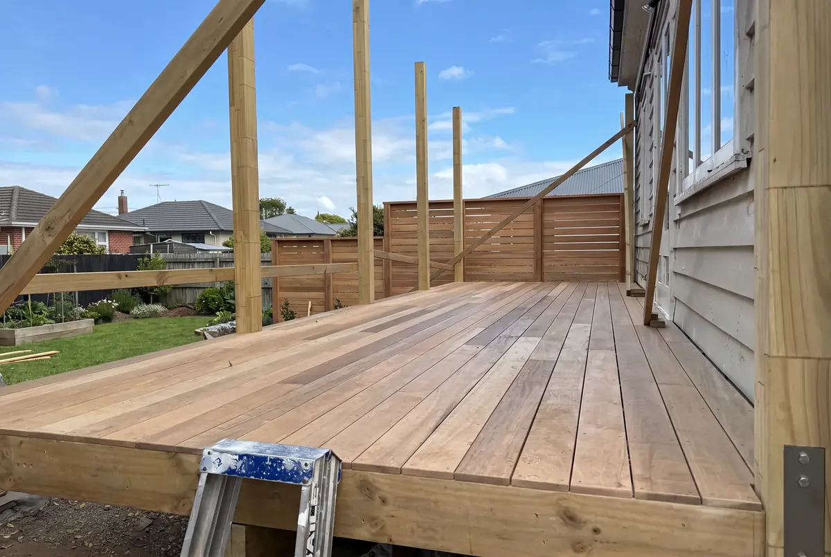 A hardwood deck being built against a weatherboard house, with the pergola posts up and a new slatted fence behind