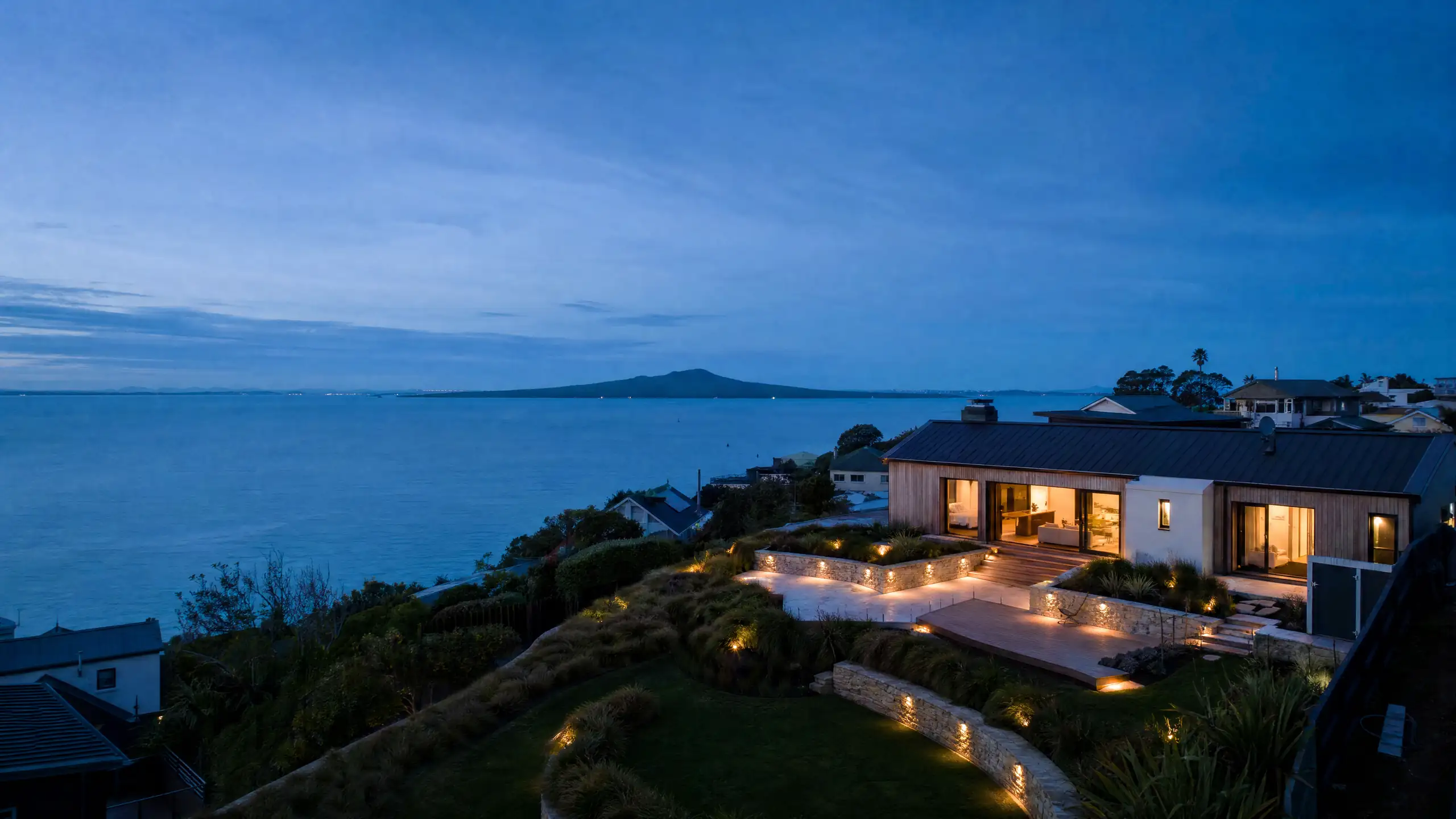 A renovated Auckland clifftop home at dusk, with a lit stone terrace and deck and Rangitoto on the horizon